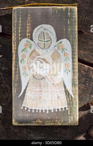 Icône en bois rustique peinte à la main représentant un ange stylisé avec des ailes florales, reposant sur une surface en bois altérée à la lumière naturelle du soleil. Banque D'Images