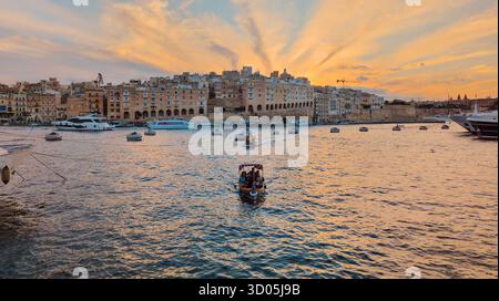 Coucher de soleil chaud sur le front de mer de Senglea sur Grand Harbour, bateaux et yachts sur l'eau ondulée, nuages pastel et raies au-dessus des bâtiments historiques en pierre : Birgu, Banque D'Images