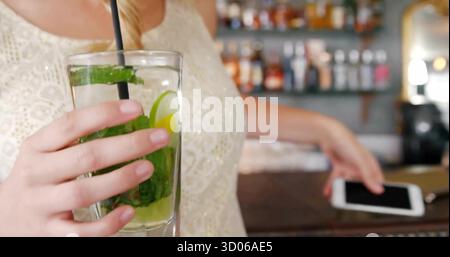 Femme portant le haut en dentelle tenant le verre de cocktail et atteignant pour smartphone au bar derrière des bouteilles Banque D'Images