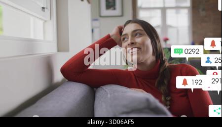 Regardant vers le haut femme portant un col roulé rouge couché sur un canapé par la fenêtre, avec des icônes de médias sociaux Banque D'Images