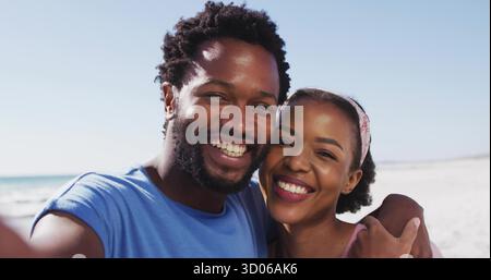 Couple afro-américain souriant prenant selfie sur la plage, avec smartphone et vêtements décontractés Banque D'Images