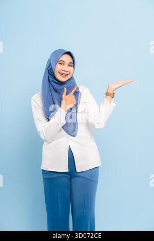 Portrait d'un jeune médecin musulman asiatique ou d'une infirmière présentant les mains ouvertes, montrant l'espace de copie ou quelque chose d'important sur un fond bleu. Banque D'Images