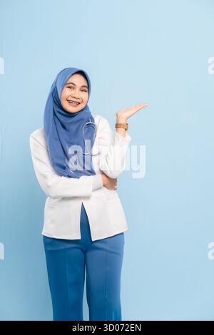 Portrait d'un jeune médecin musulman asiatique ou d'une infirmière présentant les mains ouvertes, montrant l'espace de copie ou quelque chose d'important sur un fond bleu. Banque D'Images