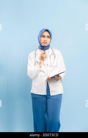 Portrait d'une jeune médecin musulmane asiatique ou infirmière tenant une tablette, levant les yeux et reposant un stylet près de son temple dans une pose réfléchie. Banque D'Images
