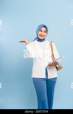 Souriante et confiante jeune étudiante en médecine ou médecin en Hijab pointant vers quelque chose d'important, isolé sur fond bleu. Banque D'Images