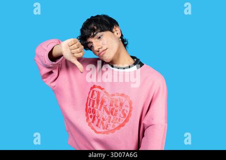 Jeune homme portant un sweat-shirt rose et donnant un geste du pouce vers le bas sur fond bleu Banque D'Images