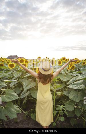 Une fille vêtiée d'une robe jaune et d'un chapeau de paille, main vers le haut, se tient avec son dos et tient un bouquet de tournesols sur un grand champ de tournesols. Un beau Banque D'Images