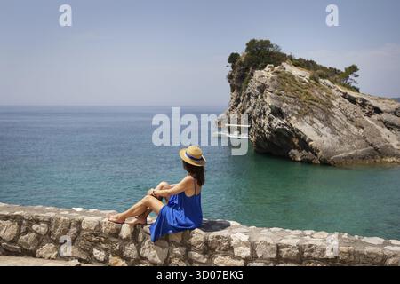 La fille, vêchée d'une robe bleue et d'un chapeau de paille, est assise près de la mer sur un rocher, sur une falaise et regarde la mer. Vacances, voyages, tourisme Banque D'Images