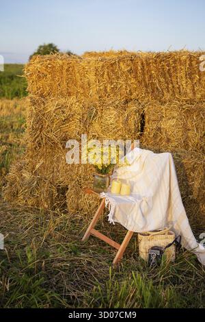 Pique-niquez dans le champ près des balles de paille. Le soleil couchant. Style rustique - chaise en bois, écossais, bouquet de fleurs, bougies Banque D'Images