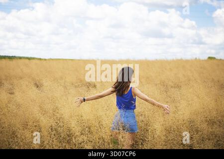 Portrait d'une jeune belle femme brune dans un t-shirt bleu et un short en denim se tient au milieu du champ, exposant son visage au soleil sur le Banque D'Images