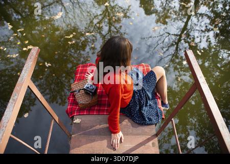 Une jeune femme assise avec son dos sur un pont près d'un lac sur une journée ensoleillée. Plaid, appareil photo, suitcase Banque D'Images
