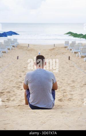 Jeune homme assis avec le dos sur une plage de sable Banque D'Images