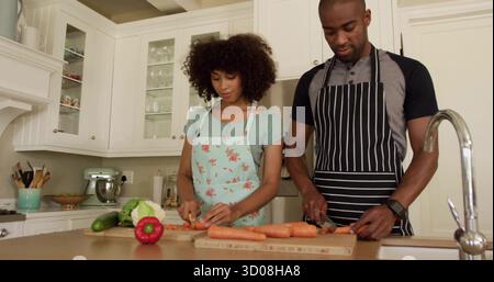 Couple hacheur portant des tabliers tranchant des carottes et des poivrons sur l'îlot de cuisine, avec des planches en bois Banque D'Images
