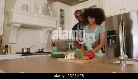 Tranchant femme dans le tablier avec couteau coupant les légumes sur l'îlot de cuisine, avec l'homme embrassant derrière Banque D'Images