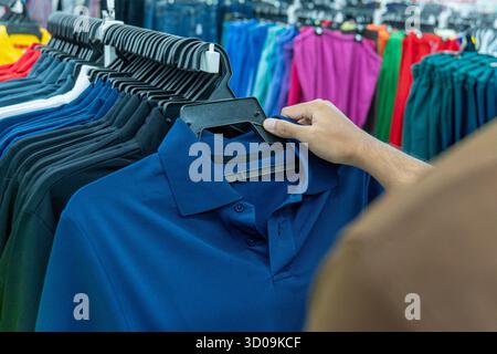 Gros plan d'une personne sélectionnant un polo bleu dans un porte-vêtements dans un magasin. Mode, shopping, vente au détail et concept de choix du consommateur. Banque D'Images