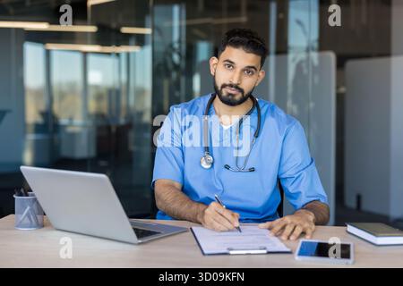 Jeune médecin masculin ou stagiaire en médecine portant des gommages bleus et un stéthoscope, assis à un bureau moderne, écrivant sur un presse-papiers avec un ordinateur portable à proximité Banque D'Images