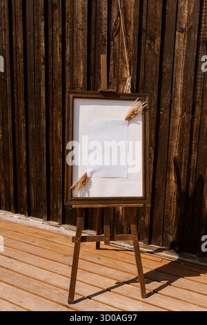 Un charmant chevalet en bois affiche un papier blanc vierge encadré contre un mur de grange brun altéré, accentué d'une délicate herbe de pampas. idéal pour le mariage Banque D'Images