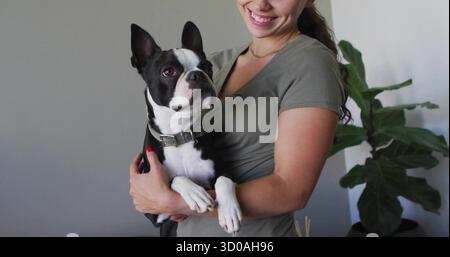 Femme souriante vêtue de façon décontractée tenant un chien Boston terrier à la maison, avec une plante de figues à feuilles de violon. Portrait, propriétaire d'animal de compagnie, moderne, confortable, style de vie, verdure, Banque D'Images