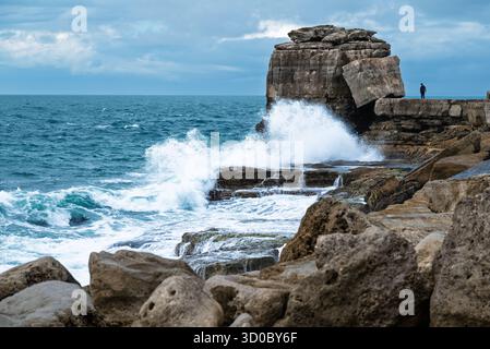 Des vagues dramatiques s'écrasent contre un affleurement rocheux alors qu'une personne se tient sur le bord de la falaise. Banque D'Images