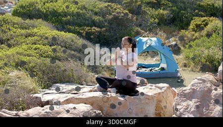 Femme méditante portant des vêtements d'activité sur rocher au camping avec tente dôme bleu clair et smartwatch Banque D'Images
