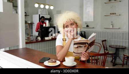 Femme de lecture portant une robe jaune penchée la tête sur la main au comptoir du café, avec livre et cappuccino Banque D'Images