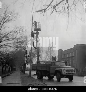 Les ouvriers soviétiques installent des isolateurs haute tension sur un poteau de service public à l'extérieur de l'armature-Insulator Plant (AIZ) à Sloviansk le 20 avril 1974. Cette photo d'archives en noir et blanc de l'URSS, la RSS d'Ukraine, montre une scène de travail industriel. Une équipe de linemen travaille sur le poteau, l'un dans le panier d'un camion de cueillette de cerises ZIL-157 (ZIL-157), tandis que d'autres grimpent sur le poteau. Cette image capture l'installation des propres produits de l'usine et la vie industrielle des années 1970 Un document précieux de la vie industrielle pacifique dans le Donbass avant la guerre. Banque D'Images