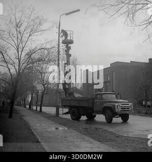 Les ouvriers soviétiques installent des isolateurs haute tension sur un poteau de service public à l'extérieur de l'armature-Insulator Plant (AIZ) à Sloviansk le 20 avril 1974. Cette photo d'archives en noir et blanc de l'URSS, la RSS d'Ukraine, montre une scène de travail industriel. Une équipe de linemen travaille sur le poteau, l'un dans le panier d'un camion de cueillette de cerises ZIL-157 (ZIL-157), tandis que d'autres grimpent sur le poteau. Cette image capture l'installation des propres produits de l'usine et la vie industrielle des années 1970 Un document précieux de la vie industrielle pacifique dans le Donbass avant la guerre. Banque D'Images