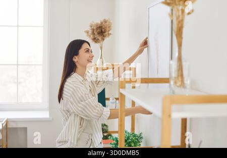 Jeune femme accrochant le cadre photo sur le mur blanc, décorant son espace de vie moderne Banque D'Images
