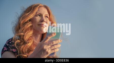 Jolie dame de gingembre en robe d'été à l'aide d'un smartphone devant le ciel Banque D'Images