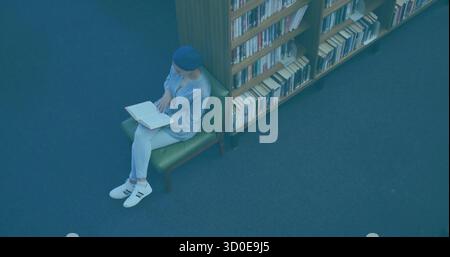 Femme du moyen-Orient lisant un livre ouvert sur une chaise verte à côté d'une étagère dans la bibliothèque, espace de copie Banque D'Images