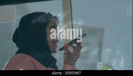 Femme parlante portant le hijab, blazer rose tenant le smartphone dans le bureau, réflexion de données, espace de copie Banque D'Images