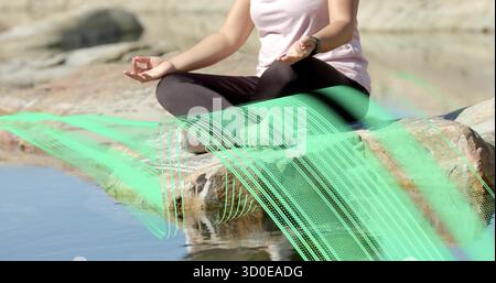 Femme en top rose, leggings méditant sur le rocher par l'eau calme, avec smartwatch, ondes numériques vertes Banque D'Images