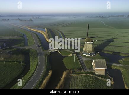 Vue aérienne d'un moulin à vent solitaire debout au milieu du paysage plat et verdoyant, projetant une longue ombre sur les champs, entrecoupés par des canaux et des routes, Blesdijke, pays-Bas. Banque D'Images