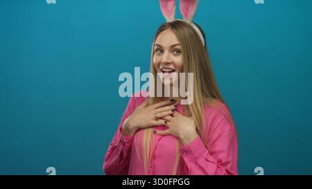 Jeune femme avec des oreilles de lapin sourit joyeusement contre un mur de fond bleu isolé, mettant en valeur des vibrations ludiques de pâques dans sa tenue rose vibrante. Banque D'Images