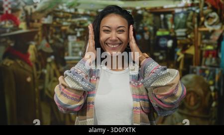 Jeune femme hispanique souriante avec les mains encadrant le visage à l'intérieur d'un bâtiment de magasin vintage rempli d'antiquités et de curiosités ; joie confiance. Banque D'Images