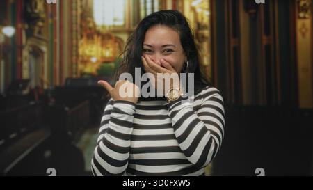 Jeune femme hispanique couvre la bouche avec la main et pointe le pouce à côté à l'intérieur de l'église ornée ; surprise amusée. Banque D'Images