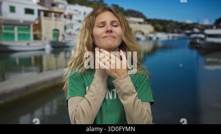 Femme serrant le cou par un port montre blonde jeune détresse de douleur dans un cadre extérieur de front de mer. Banque D'Images