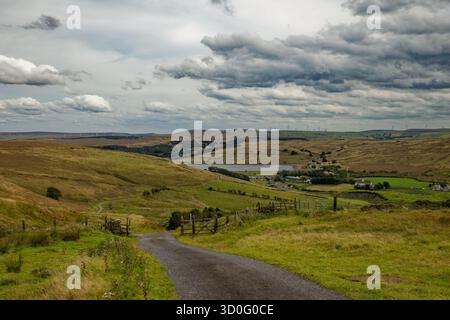 Une étroite route rurale serpente à travers un sombre paysage de landes Pennine avec un réservoir dans une vallée lointaine, Burnley, Lancashire. Banque D'Images