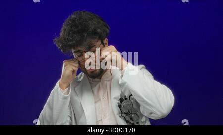 Jeune homme en blouse de laboratoire blanche avec des lunettes prétendant pleurer sur un fond bleu, mettant en valeur un geste humoristique et expressif. Banque D'Images