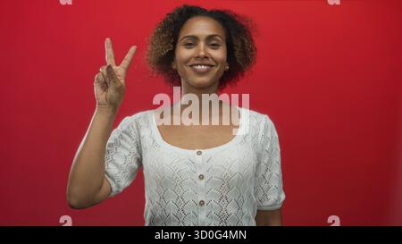 Femme pointant l'index et montrant un signe de paix à deux doigts dans un studio avec fond rouge, portant un cardigan en tricot blanc et souriant ; pois de confiance Banque D'Images