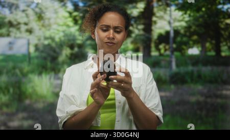 Femme tenant un avocat meurtri, la main sur le menton, le sourcil sillonné, inspectant les fruits dans le parc près des arbres ; pensée confuse. Banque D'Images
