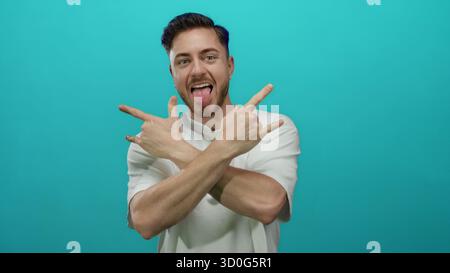 Jeune homme avec la barbe faisant des gestes de main rock sourit avec confiance sur un fond bleu vif, mettant en valeur l'énergie et la jeunesse. Banque D'Images