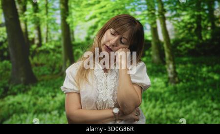 Jeune femme bâillant la main sur la bouche et les yeux fermés, portant un chemisier blanc et une montre-bracelet dans un cadre forestier ; somnolence repos reflet. Banque D'Images