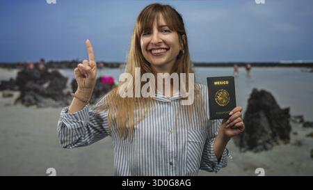 Femme souriante tient le passeport mexicain dans la main droite et pointe l'index vers le haut tout en se tenant debout sur une plage de sable avec des roches volcaniques et l'horizon de la mer ; trav Banque D'Images