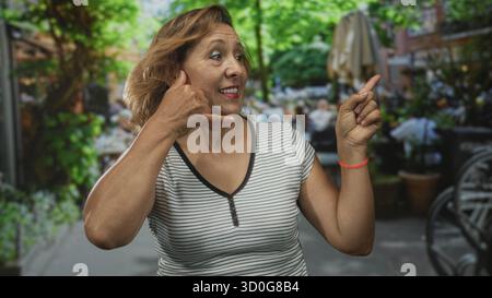 Femme montre Call me geste avec la main et pointe doigt à la caméra sur la terrasse de la rue restaurant sous la canopée verte ; invitation joyeuse. Banque D'Images