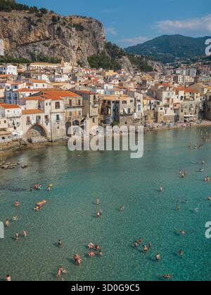 Vue aérienne des toits en terre cuite baignés de soleil en cascade vers la mer Azur, où les nageurs parsèment les eaux cristallines près de la cathédrale historique de Cefalù, Cefalù, Sicile, Italie. Banque D'Images