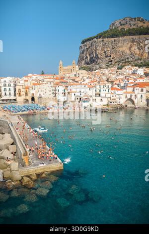 Vue aérienne de nageurs baignés par le soleil parsemant les eaux turquoises sous la cathédrale historique de Cefalù et Rocca di Cefalù, Cefalù, Sicile, Italie. Banque D'Images