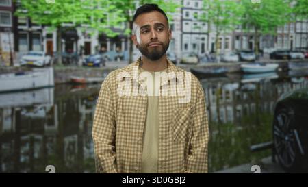 Jeune homme hispanique debout près de l'eau du canal dans la rue à amsterdam avec le visage pensif et les mains sur les côtés ; contemplation tranquille. Banque D'Images