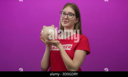 Jeune femme blonde portant une chemise de bénévole souriant tient une tirelire en studio ; générosité don de charité. Banque D'Images