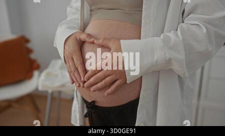 Femme enceinte avec les mains sur le ventre se tient à l'intérieur, transmettant l'anticipation de la maternité dans un cadre de maison confortable avec un éclairage doux. Banque D'Images